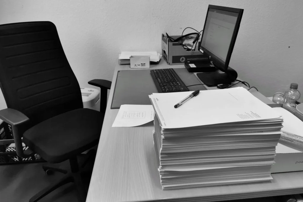 Black and white image of a cluttered office desk with a computer, chair, and paperwork